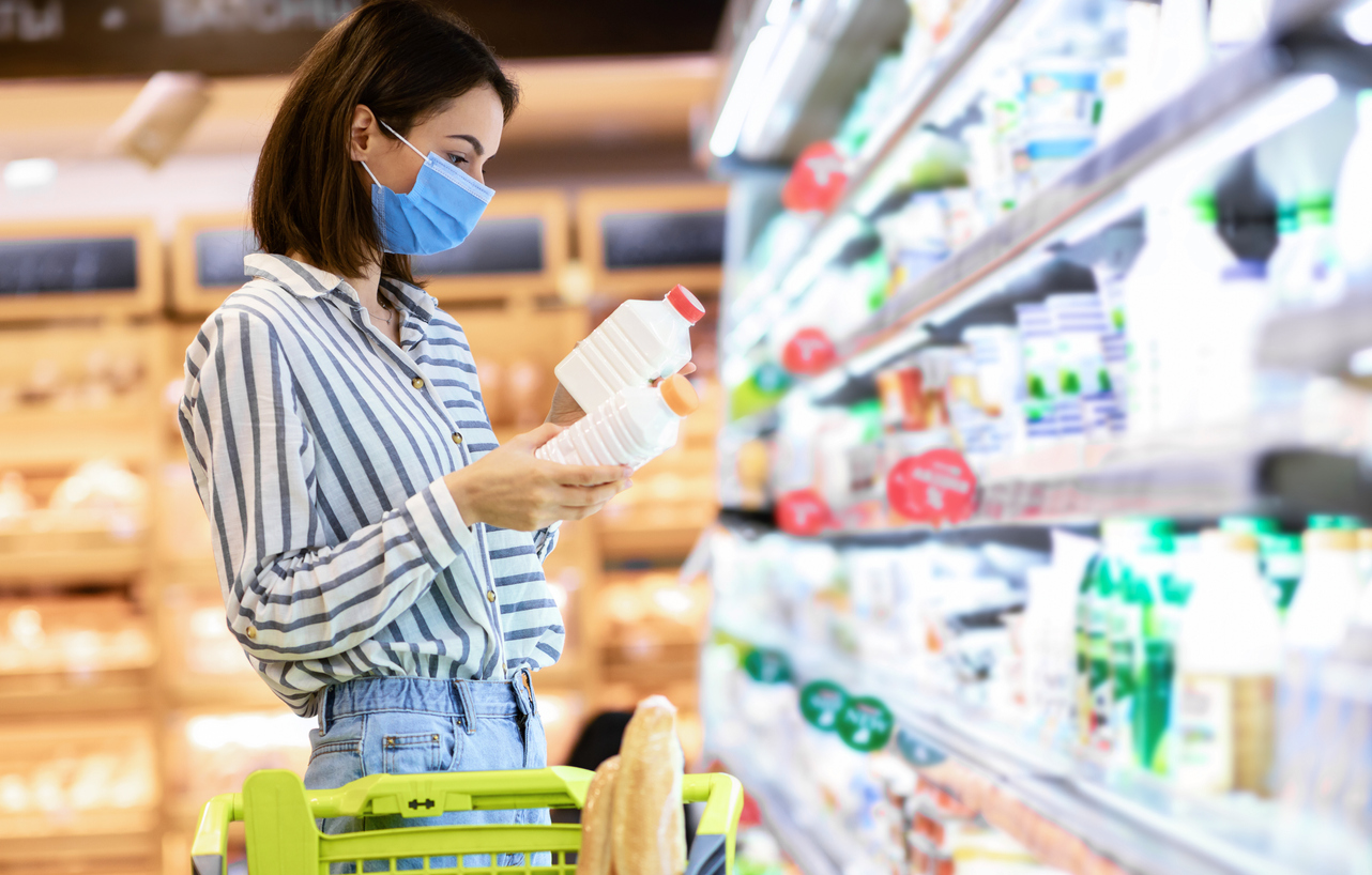 Young woman in medical disposable mask holding two bottles of milk or yoghurt, standing near the freezer, choosing dairy products, making decisions, checking expiry date